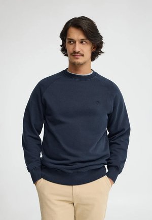 Uomo con capelli scuri di media lunghezza, indossa una felpa blu navy e pantaloni beige, in piedi con le mani nelle tasche, guarda di lato.