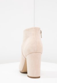 Bota de tobillo de ante beige con punta redonda, tacón alto y cuadrado, y detalle de cremallera lateral. Textura suave y diseño minimalista.