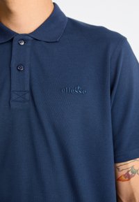 Tamsiai mėlynas polo marškinėliai su tekstūruotu audiniu, trimis mygtukais ir siuvinėtu logotipu ant krūtinės. Matomos trumpomis rankovėmis.