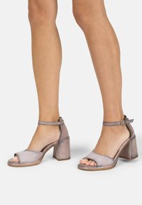 Open-toe block-heeled sandals in a soft, metallic lavender material with ankle strap and buckle detail. Smooth texture, minimalistic design.