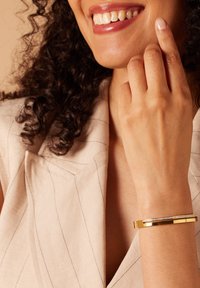 Gouden armband met een gladde metalen afwerking, voorzien van een rij heldere edelstenen en een rechthoekige vorm, gedragen aan een hand met subtiele huidtinten.