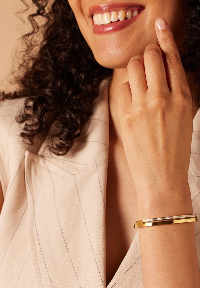 Gouden armband met een gladde metalen afwerking, voorzien van een rij heldere edelstenen en een rechthoekige vorm, gedragen aan een hand met subtiele huidtinten.
