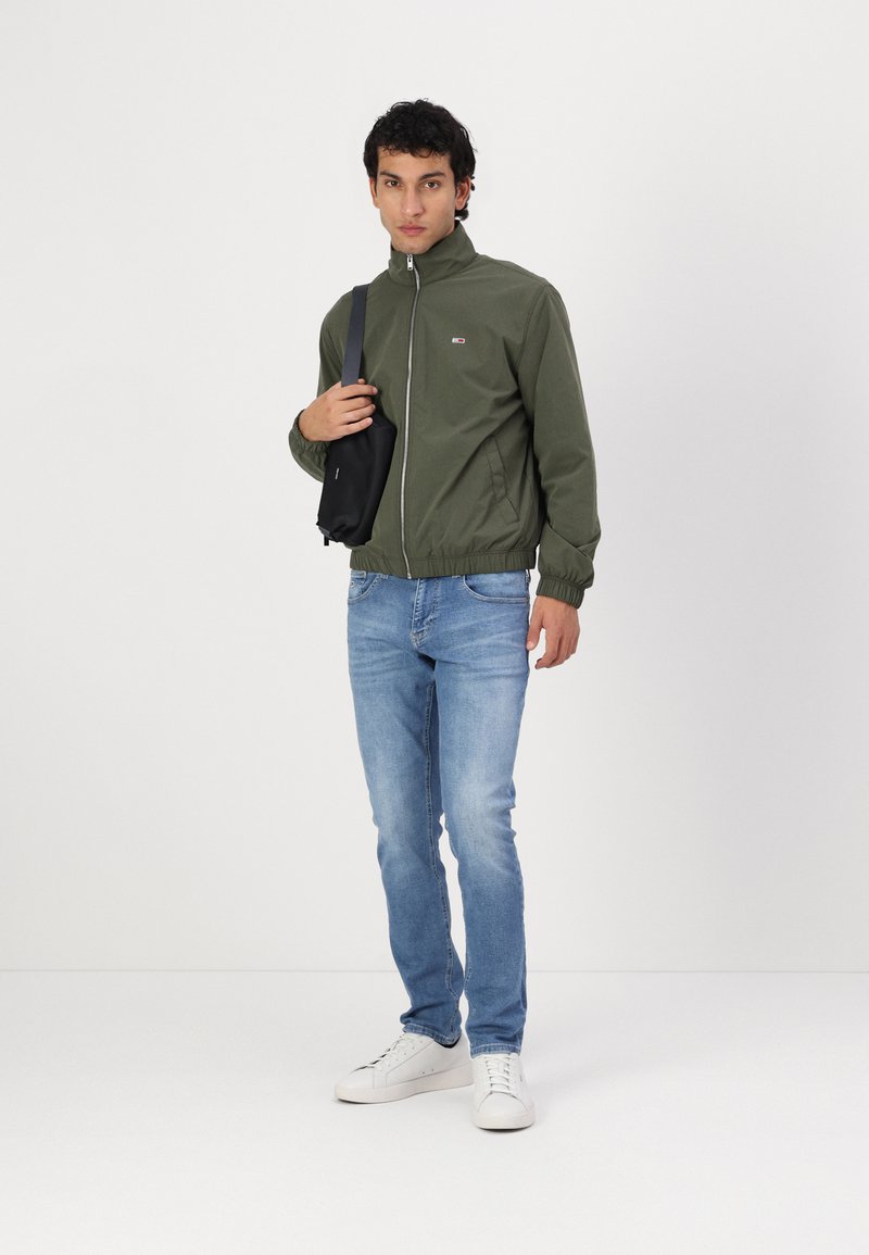 Veste zippée verte avec col montant, jeans en denim et baskets blanches. Le modèle tient un petit sac à bandoulière noir. Texture de tissu lisse.