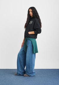 Fekete kapucnis pulóver elülső zsebbel és grafikai tervezéssel, lazán szabott, világoskék farmernadrággal és a derékon megkötött zöld inggel párosítva.