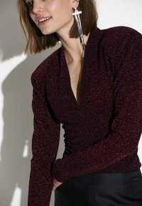 Bordeaux glinsterende top met lange mouwen en een v-hals ontwerp. Strak aansluitend met geplooide details bij de taille. Zilveren geometrische oorbellen.