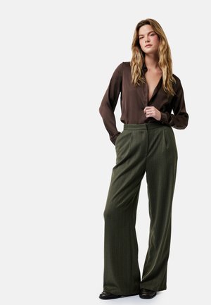 Frau mit langen blonden Haaren trägt eine dunkelbraune Seidenbluse und hoch taillierte, weit geschnittene olivgrüne Hose, steht mit einer Hand in der Tasche.