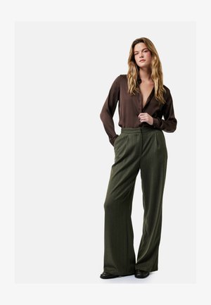 Mujer con cabello largo y rubio lleva blusa de seda marrón oscuro y pantalones anchos de pierna alta de color verde oliva, de pie con una mano en el bolsillo.