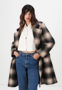 Kariertes Wollmantel in Beige- und Schwarztönen, mit langen Ärmeln und breiten Revers, getragen über einem weißen cropped Shirt und blauen Jeans mit lockerer Passform.