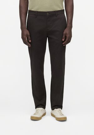 Mann trägt schwarze Hose mit geradem Bein, beige Sneakers mit Gummisohlen und ein olivgrünes Hemd, steht vor weißem Hintergrund.