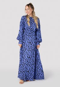 Robe maxi en tissu bleu avec un motif de pois noirs, dotée de longues manches bouffantes, d'une taille élastique et d'une fente sur le côté pour plus de facilité de mouvement.