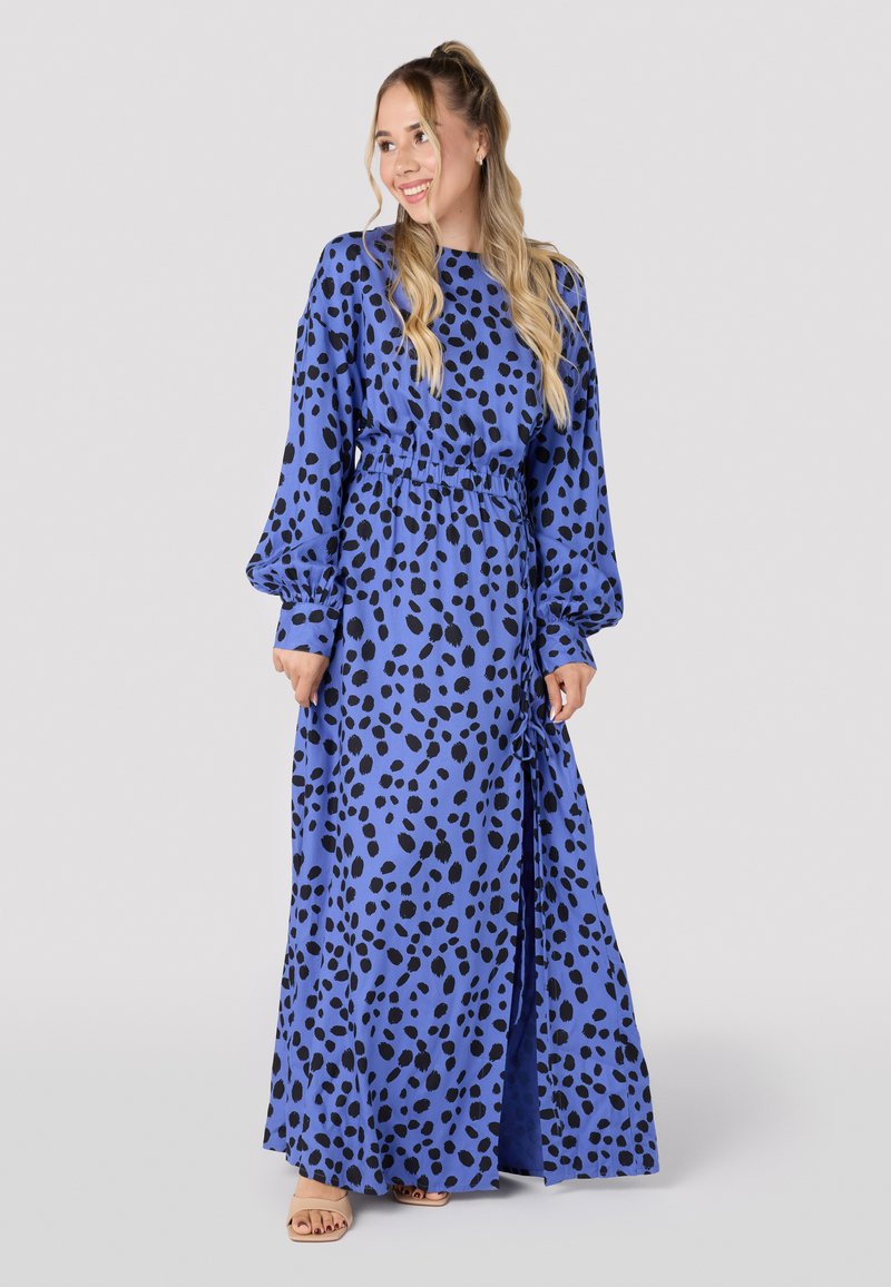 Robe maxi en tissu bleu avec un motif de pois noirs, dotée de longues manches bouffantes, d'une taille élastique et d'une fente sur le côté pour plus de facilité de mouvement.