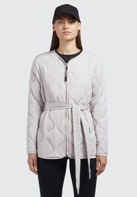 Polstret jakke i lys grå med quilted bølgermønster, lynlås foran og matchende bælt. Båret med en sort kasket og mørk overdel under.