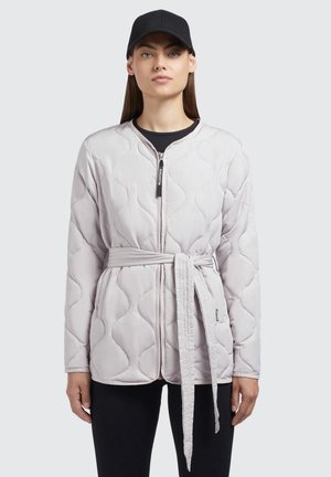 Giacca imbottita di grigio chiaro con motivo a onde trapuntato, chiusura con zip e cintura abbinata. Indossata con un berretto nero e una maglietta scura sotto.