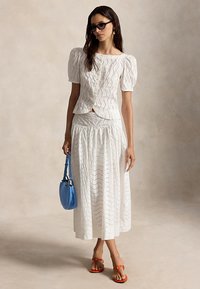 Γυναίκα που φοράει λευκό μπλουζάκι με φουσκωτά μανίκια από σκαλίσματα και ταιριαστή φούστα, κρατώντας μπλε τσάντα χειρός, φοράει σκούρα γυαλιά ηλίου και πορτοκαλί σανδάλια.