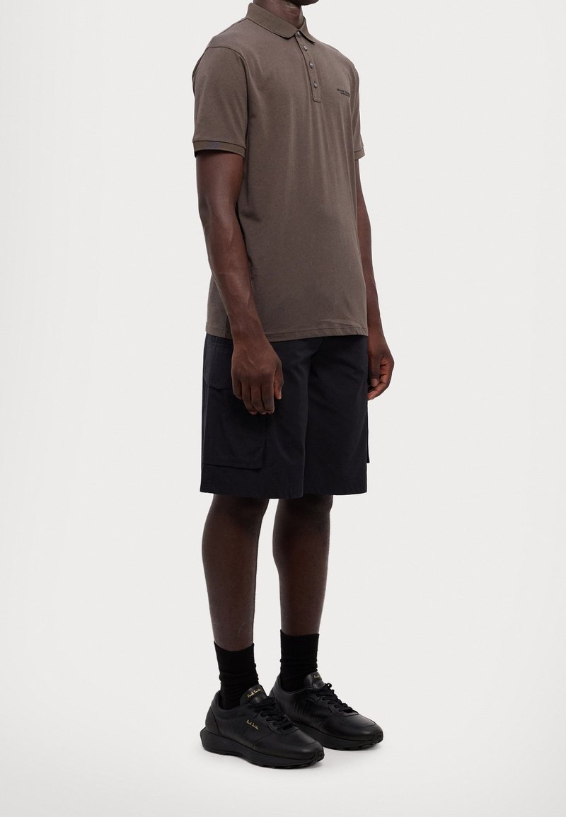 Homme portant un polo marron à manches courtes, un short cargo noir, des chaussettes noires et des baskets noires sur fond blanc uni.