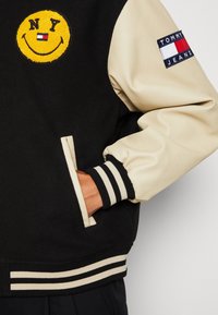 Černá vlněná a krémová kožená varsity bunda s žlutou nášivkou úsměvu a nápisem "NY", pruhovanými žebrovanými manžetami a logem Tommy Jeans na rukávu.