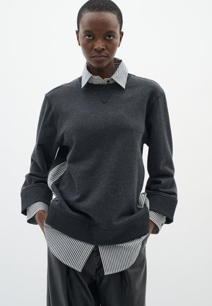 Felpa grigia con scollo rotondo, indossata sopra una maglietta a righe bianco e nero. I polsini sono leggermente oversized e il tessuto appare morbido.
