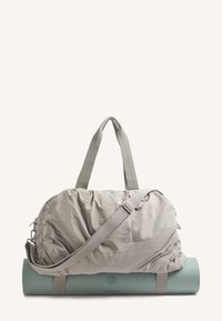 Vaaleanharmaa, pöhöttyvä duffelilaukku, jossa on lyhyet kahvat ja pitkä olan yli kulkeva hihna, lepäämässä teksturoidun mintunvihreän joogamaton päällä.