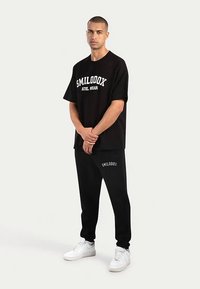 Czarny bawełniany t-shirt z białym napisem "SMILODOX", w zestawieniu z czarnymi dresami z tym samym logo; model nosi białe sneakersy.