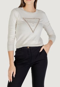 Top blanco de manga larga con un logotipo triangular de "GUESS" en el centro, decorado con strass, combinado con pantalones negros.