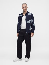 Homme debout portant des lunettes transparentes, chemise bleue marine à fleurs par-dessus un haut blanc, pantalon noir et baskets blanches sur un fond clair uni.