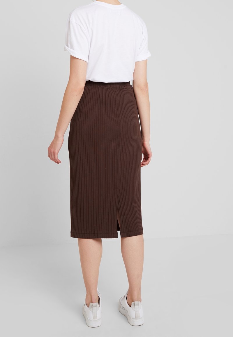 Brauner, gerippter Midirock mit einem Schlitz hinten, ausgestattet mit einem dehnbaren Bund. Kombiniert mit einem weißen T-Shirt und weißen Sneakers.