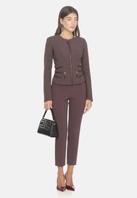 Giacca aderente color bordeaux con chiusura a zip e due tasche con zip, abbinata a pantaloni su misura dello stesso colore. Gli accessori includono una borsa nera.