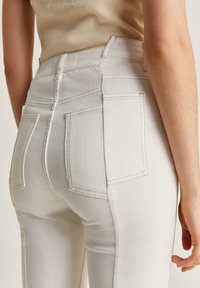 Jeans blancs taille haute avec une texture lisse, présentant des poches arrière et des coutures contrastantes. La coupe met en valeur une silhouette ajustée.