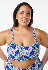 Soutien-gorge de sport à imprimé floral dans des tons de violet, bleu et jaune. Doté de larges bretelles et d'un décolleté en cœur. Tissu extensible et texturé.