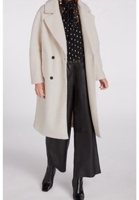 Hvid tekstureret frakke med et langt, lige design, der har to sorte knapper, båret over en sort mønstret bluse og sorte læderbukser.
