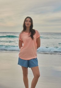 Persika värvi, lühikeste varrukatega T-särk ümarate servadega, koos siniste triibuliste lühikeste pükste ja ookeani lainetega taustal rannal.