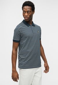 Νεαρός άνδρας που φοράει μπλε σκούρο και λευκό ριγέ πόλο μπλουζάκι και ανοιχτόχρωμο παντελόνι, κοιτάζοντας προς τα αριστερά του μπροστά σε απλό λευκό φόντο.