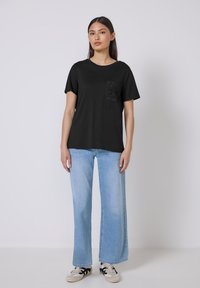 Sort t-shirt med korte ærmer og en struktureret firkantet lomme, parret med lyseblå vide jeans og sorte sko, opstillet mod en ensfarvet baggrund.