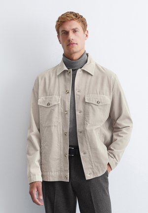 Mann in beiger Cordjacke über einem grauen Rollkragenpullover und dunklen Hosen, der vor einer schlichten weißen Wand steht.