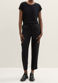 Schwarzes kurzärmliges Oberteil, kombiniert mit schwarzen Hosen mit Zugbandtaille. Das Model trägt schwarze Oxford-Schuhe. Glatter Stoff, lockere Passform.