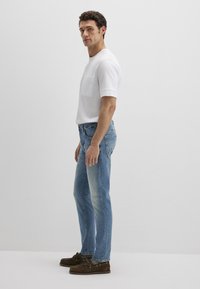 Weißes T-Shirt mit kurzen Ärmeln und Brusttasche, kombiniert mit ausgewaschenen blauen Jeans und braunen Wildleder-Loafers. Neutrale Hintergrundfarbe.