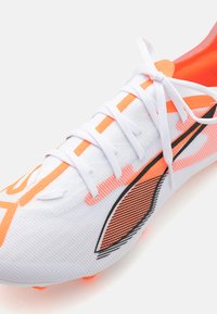White athletic shoe with orange accents, textured synthetic upper, flat laces, and black graphic details. Designed for performance.