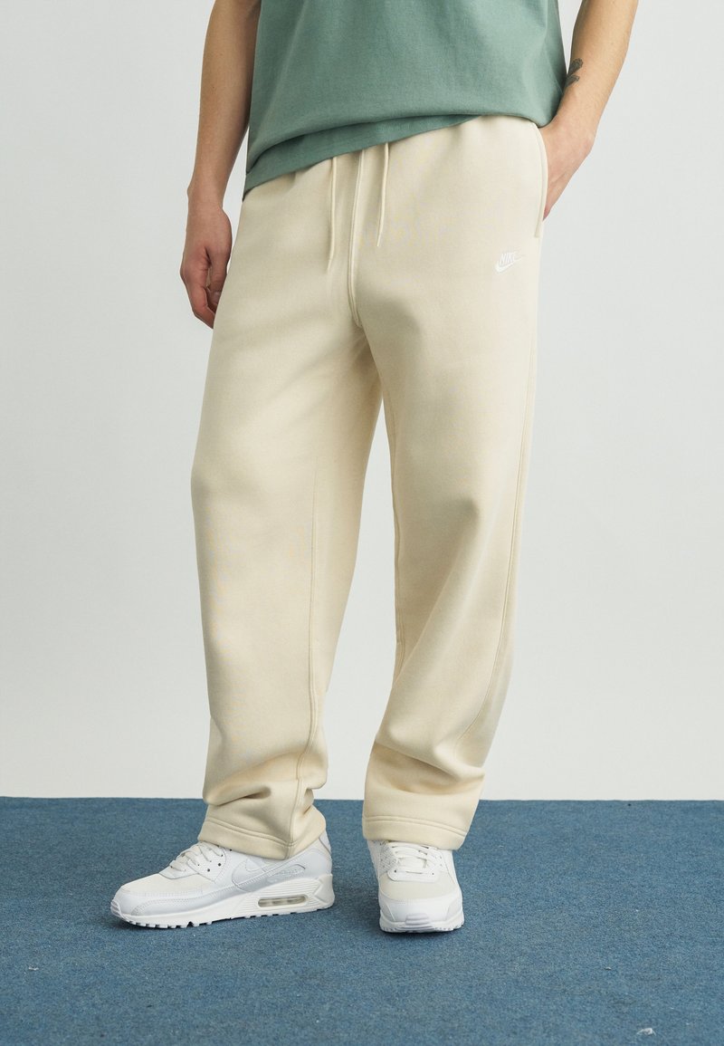 Asmuo dėvi smėlio spalvos Nike sportines kelnes ir baltus Nike kedus, stovintis ant mėlyno kilimo, rankos atpalaiduotos šonuose.