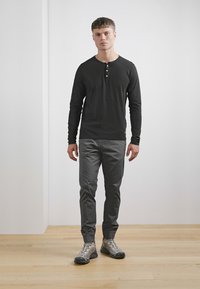 Schwarzes Langarm-Henley-Shirt mit drei Knöpfen, graue schmale Hose und graue Wanderschuhe, vor einem hellen Hintergrund auf einem Holzboden.