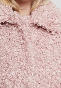 Růžový texturovaný kabát s kudrnatou tkaninou, širokým límcem a viditelnými prošitými detaily. Tkanina má měkký, nadýchaný vzhled.