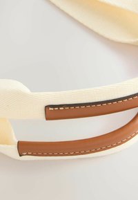 Bomullsrem i off-white med bruna läderdetaljer. Har söm längs kanten och en slät yta. Justerbar längd.