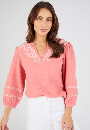 Mujer vistiendo una blusa rosa con bordado floral blanco en las mangas y el escote, combinada con pantalones blancos, posando frente a un fondo liso.