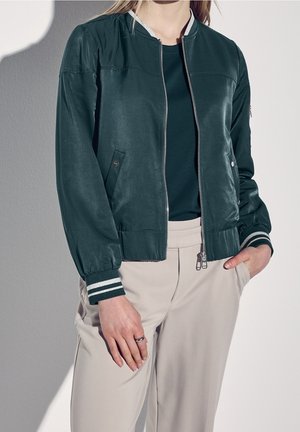 Person iført mørkegrøn bomberjakke med stribede manchetter, sort skjorte, beige bukser, stående med én hånd i lommen og den anden ned langs siden.