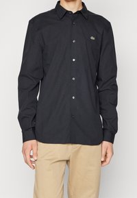 Camisa negra abotonada de algodón, con cuello clásico, mangas largas y un pequeño logotipo verde en el pecho izquierdo.