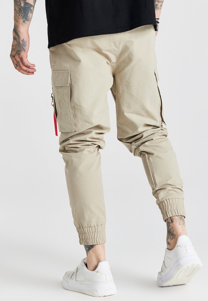 Homme portant un pantalon cargo beige de style jogger et des baskets blanches, montrant ses bras et chevilles tatoués sur un fond uni clair.