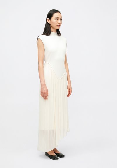 Μακριά, αμάνικη κρεμ φόρεμα με καμπυλωτό σκίσιμο και πιετέ λεπτομέρειες, λεία πάνω επιφάνεια, και απλά �μαύρα παπούτσια μπαλαρίνας.