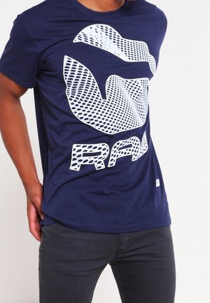 Mann trägt ein dunkelblaues T-Shirt mit großem weißem Grafikmotiv und dem Schriftzug "RAW" auf der Vorderseite, kombiniert mit einer dunklen Hose.