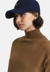 Frau mit kurzen braunen Haaren, die eine marineblaue Mütze und einen braunen gerippten Rollkragenpullover trägt und leicht nach links schaut.