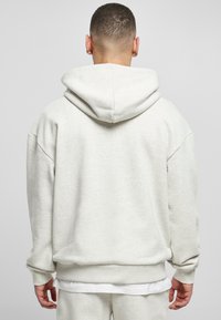 Uomo con capelli corti che indossa una felpa grigia chiara e pantaloni della tuta abbinati, visto da dietro contro uno sfondo chiaro e semplice.