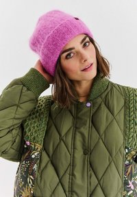 Žiarivo ružová pletená čiapka a zelená prešívaná bunda s textúrovanými rukávmi. Kvetinový vzor na boku bundy s rôznymi farbami a tvarmi.